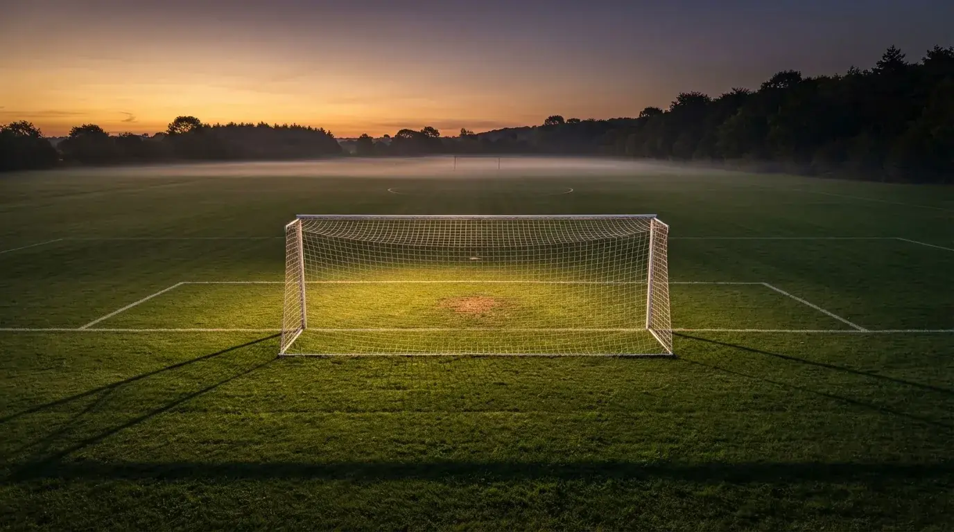 Ein Fußballtor mit Netz bei Sonnenuntergang auf einem leeren Platz, die Schatten der Torpfosten liegen lang auf dem Gras.