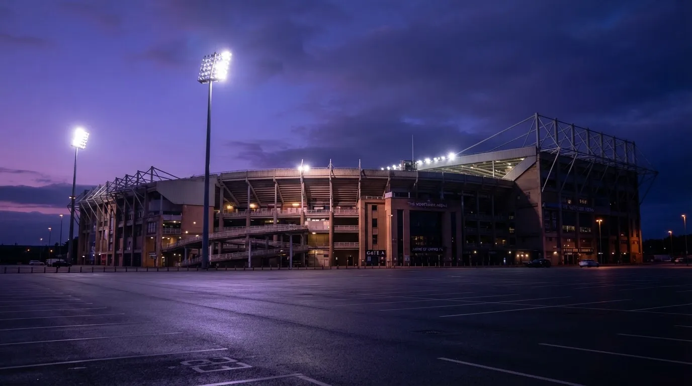 Ein Fußballstadion von außen in der Dämmerung, hohe Flutlichtmasten vor einem violetten Himmel, davor ein leerer Parkplatz.
