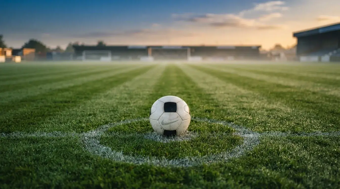 Ein Fußball liegt auf dem Anstoßpunkt eines Rasens bei Sonnenaufgang, Tau glitzert auf den Halmen.