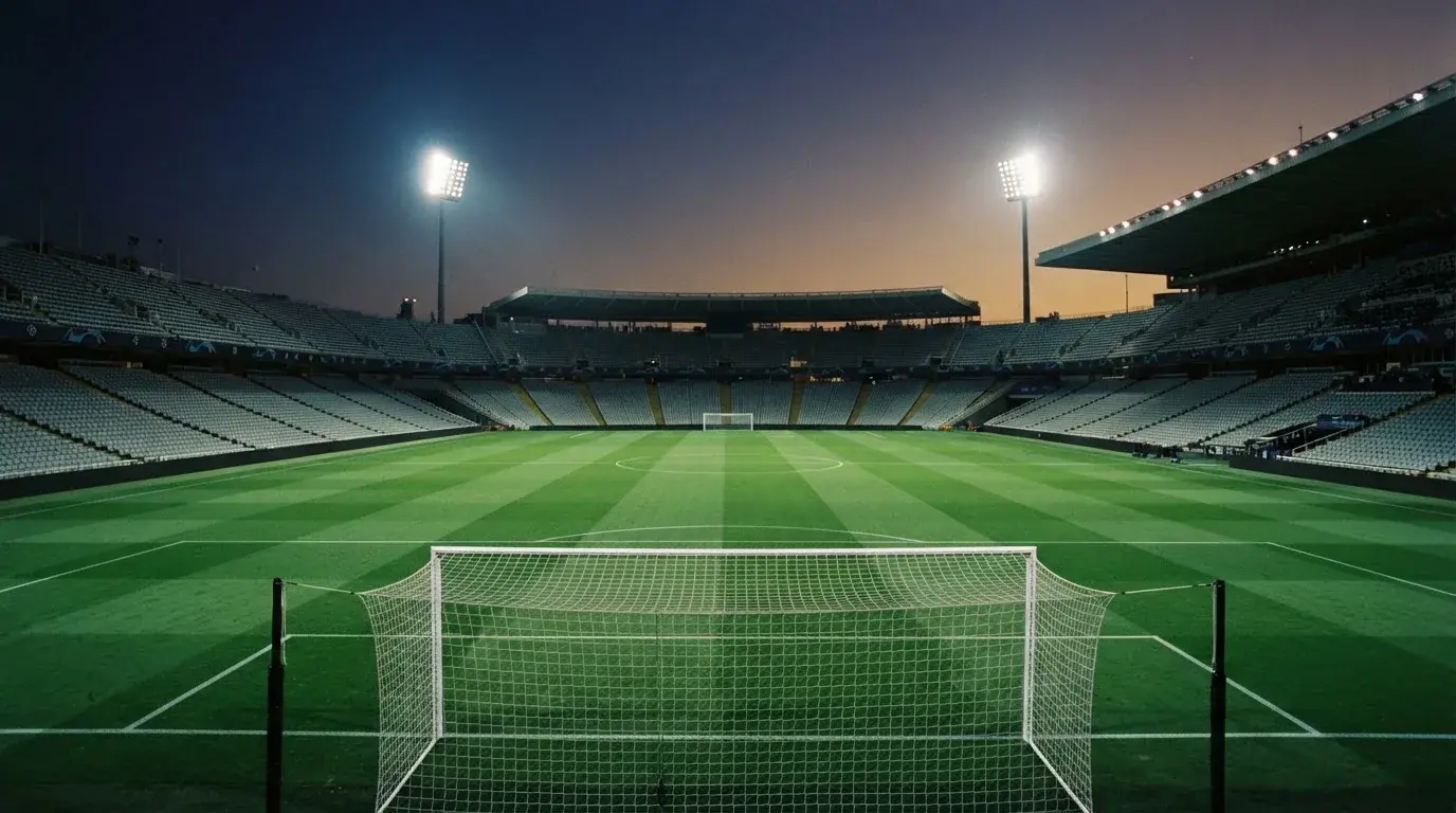 Eine leere Stadiontribüne unter Flutlicht aus der Ferne betrachtet, die Rasenfläche leuchtet grün in der Dämmerung.