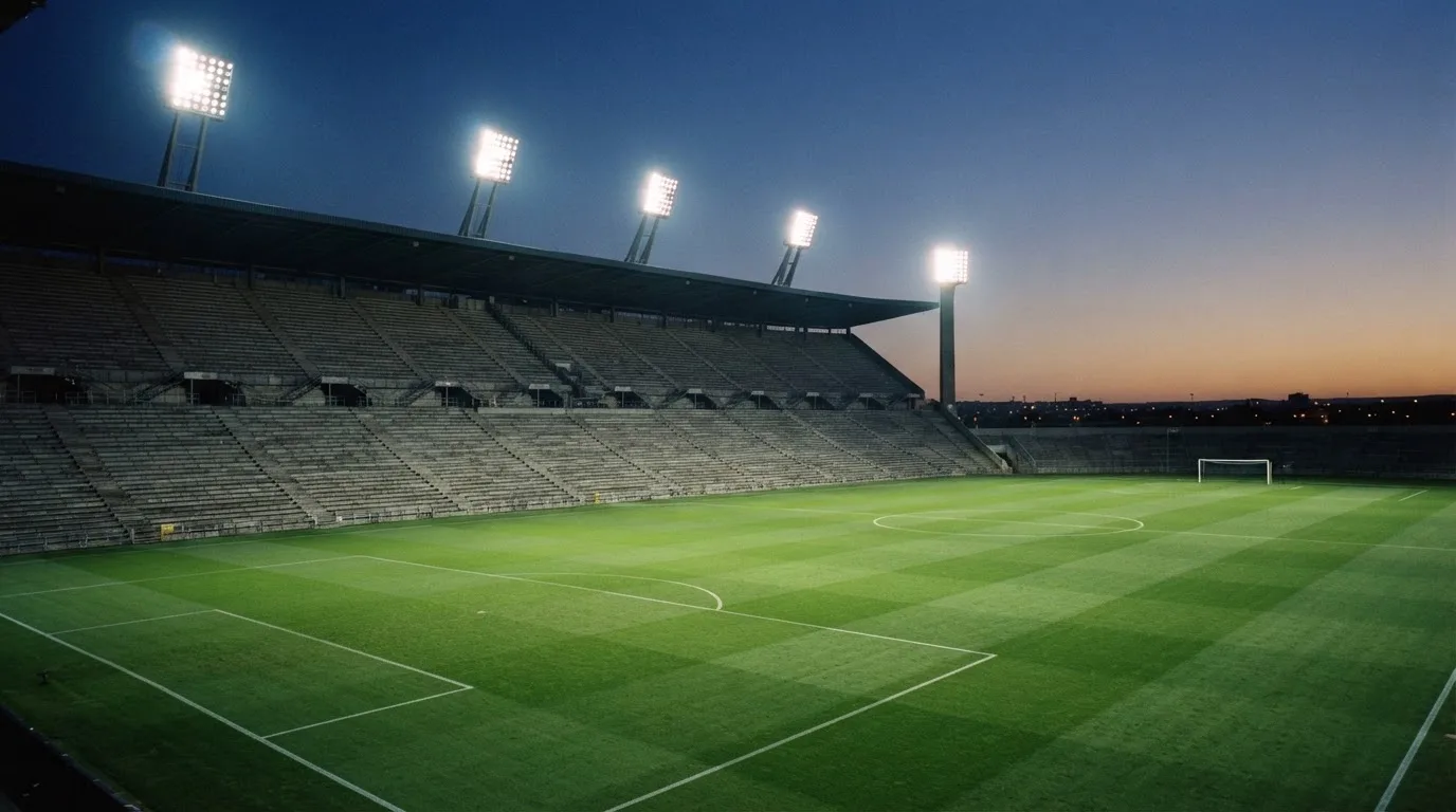 Eine leere Stadiontribüne unter Flutlicht aus der Ferne betrachtet, die Rasenfläche leuchtet grün in der Dämmerung.
