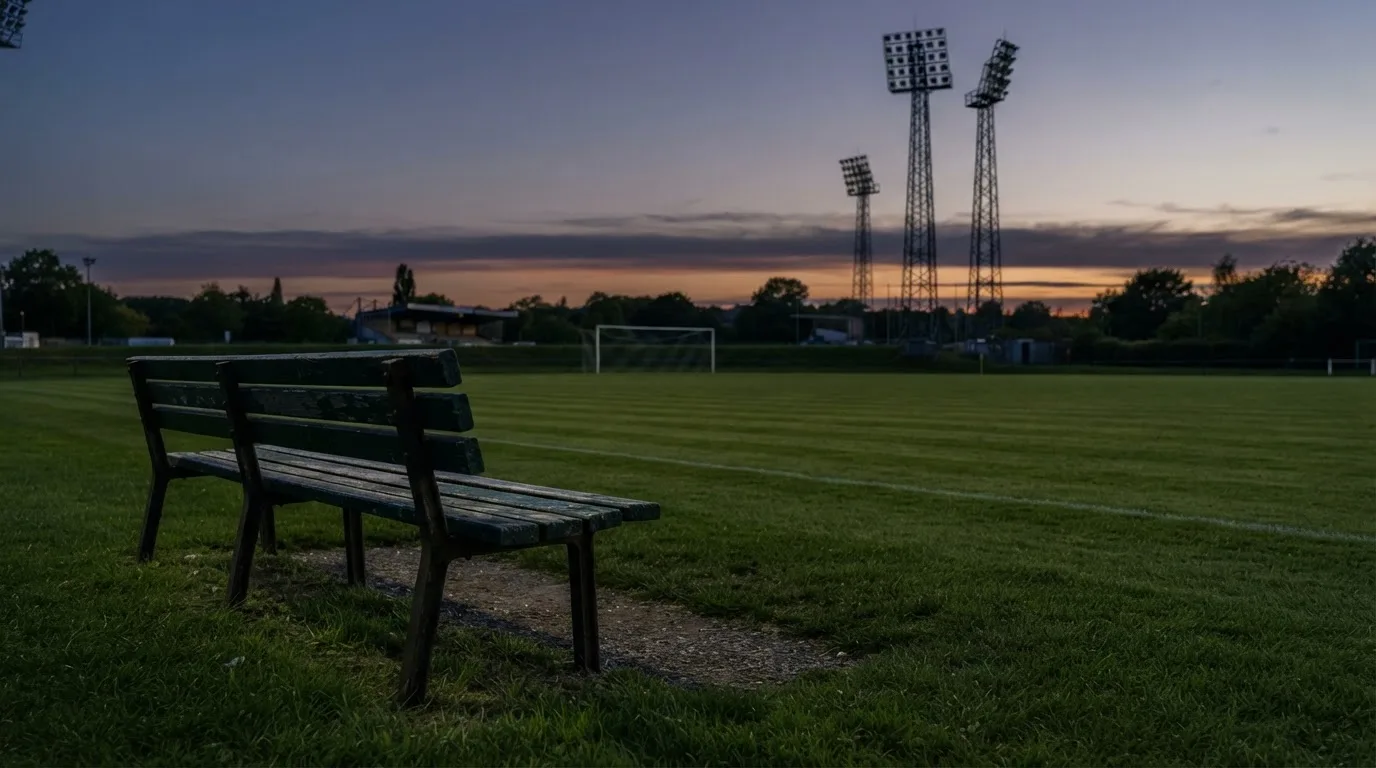 Eine leere Parkbank am Rand eines Fußballfelds in der Abenddämmerung, im Hintergrund ragen dunkle Flutlichtmasten in den Himmel.