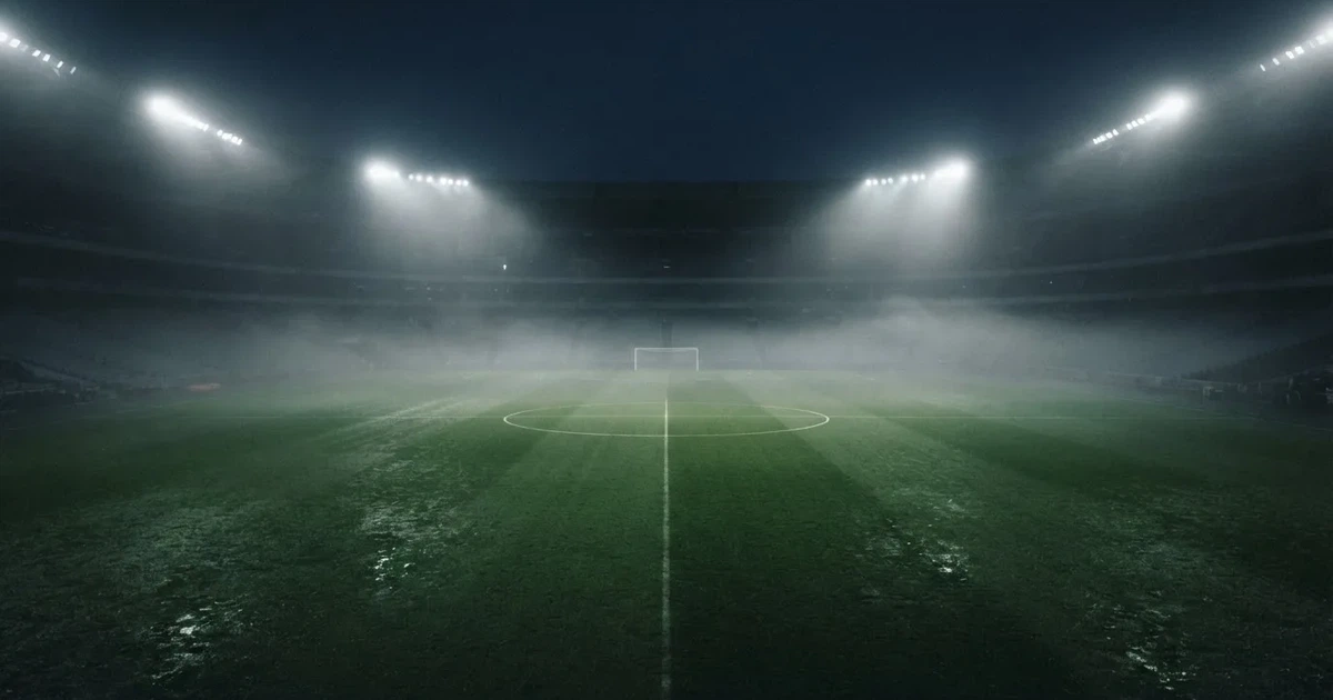 Ein Stadion unter Flutlicht, aus großer Entfernung über eine dunkle Wiese fotografiert, am Himmel zieht Nebel auf.