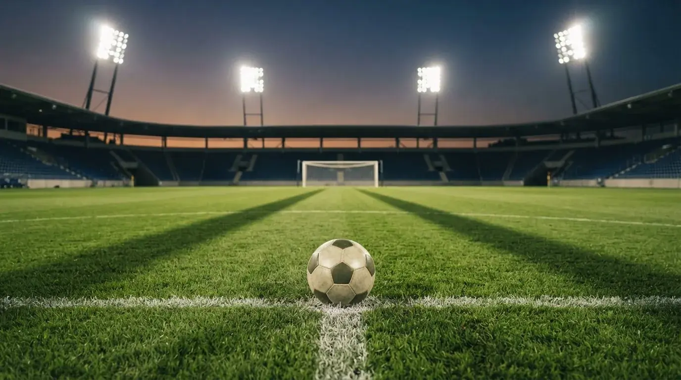 Ein Fußball liegt auf dem Anstoßpunkt eines leeren Rasenplatzes, im Hintergrund steht ein Stadion unter Flutlicht in der Dämmerung.
