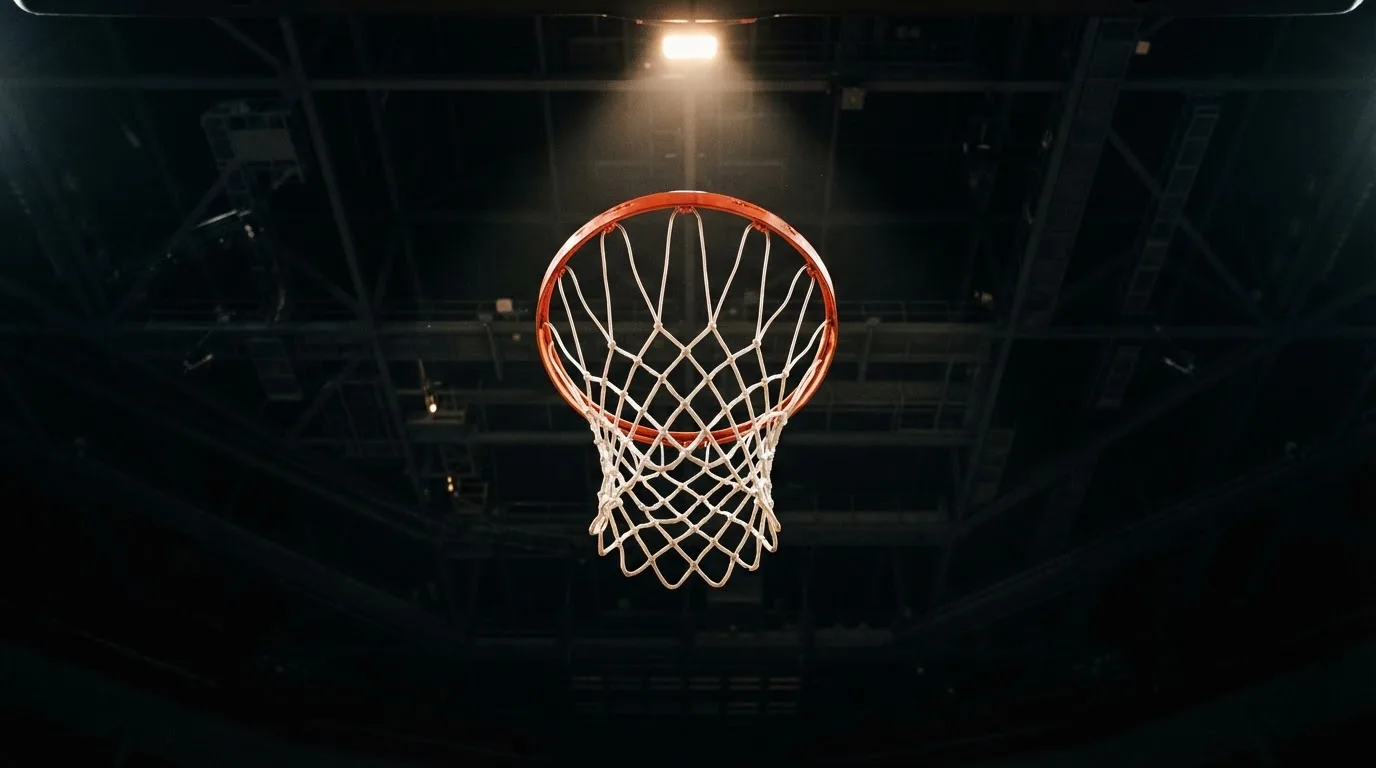 Ein Basketballkorb von unten gesehen vor dem dunklen Hallendach, das Netz hängt ruhig im Scheinwerferlicht.